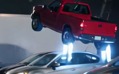A red pickup flying over a row of parked cars on blue jets at night